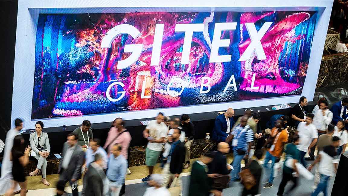 Attendees walk past a large digital screen displaying "GITEX GLOBAL" at Dubai World Trade Centre, showcasing the scale and energy of the Gulf’s leading tech event.