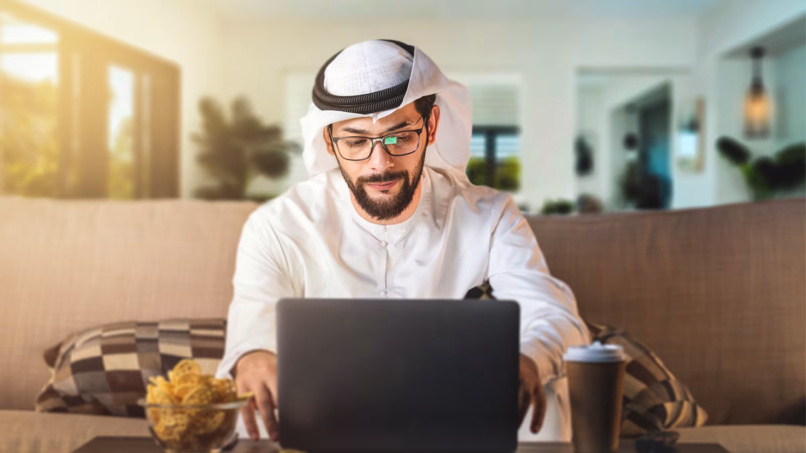 Gulf man working remotely from home on a laptop, wearing traditional attire, seated in a modern living room with natural li